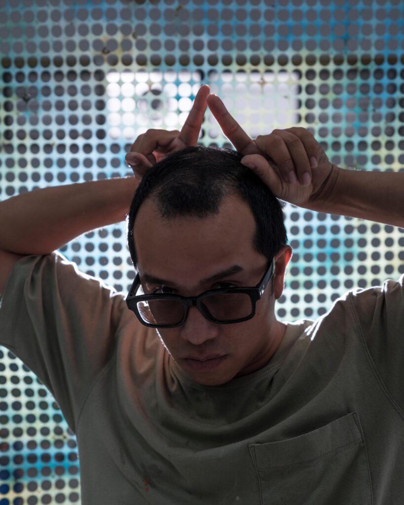 Tada Hengsapkul raising two fingers above his head in front of a dotted artwork.