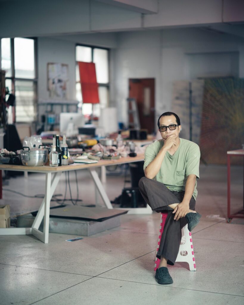Tada Hengsapkul seated on a stool with one hand over his mouth in his studio.