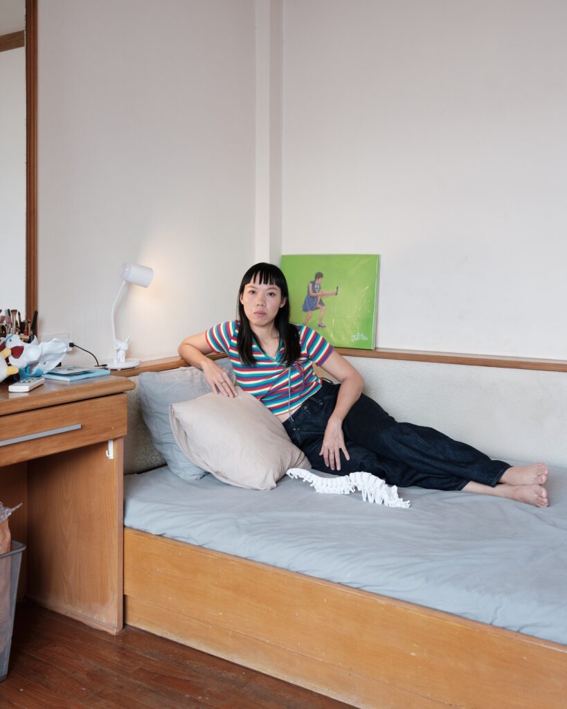 Wilawan reclining on a bed in a striped top, with anatomical models and a painting nearby.