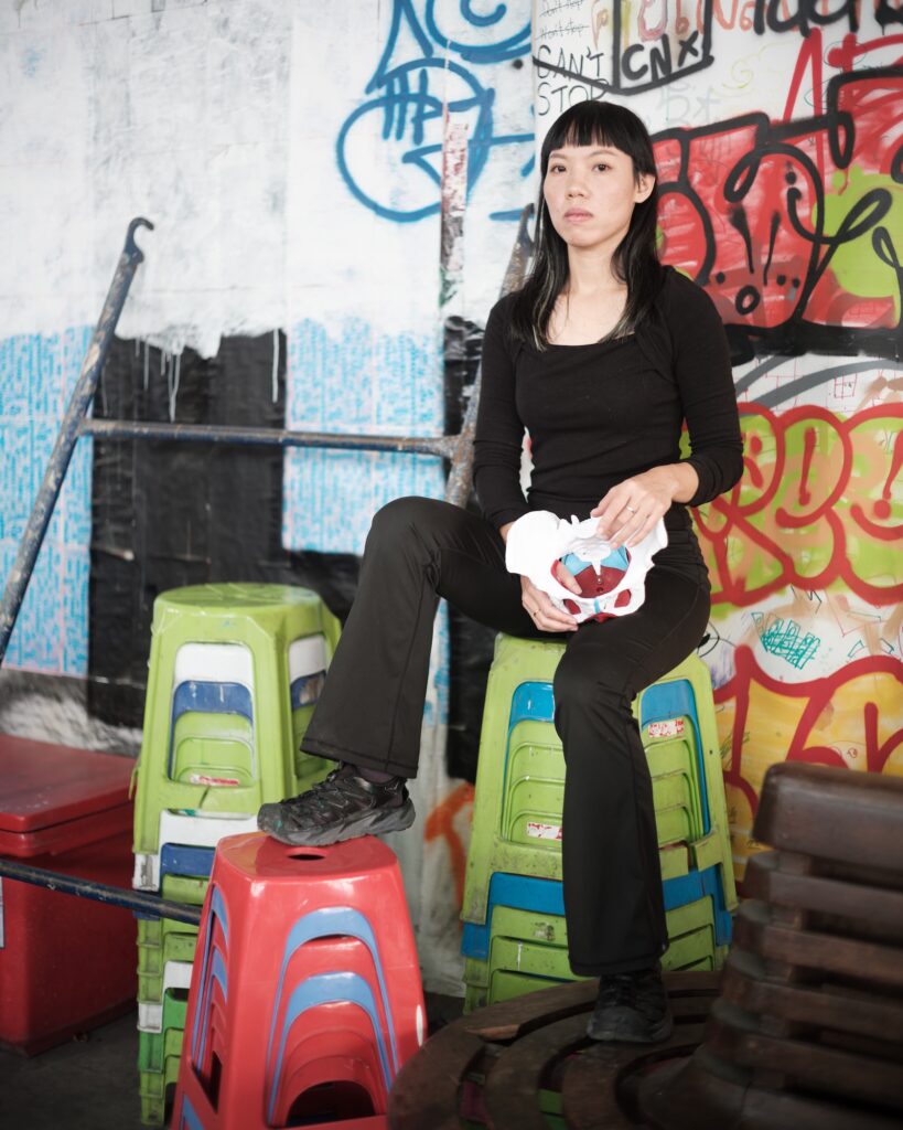 Wilawan seated on stacked colourful stools, holding an anatomical pelvis model against a graffiti-covered wall.