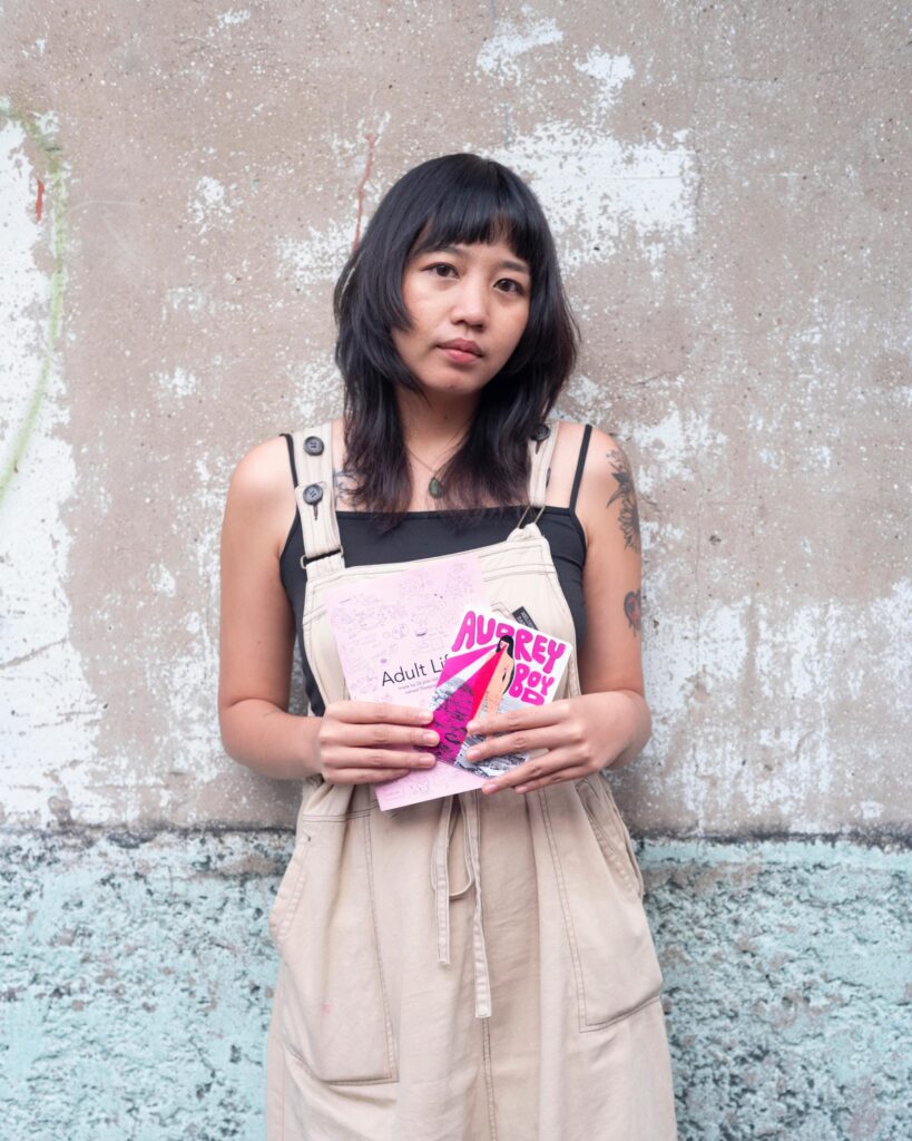 Member of Zua, Cinema and Chalan holding a pink zine against a weathered wall in Bangkok.
