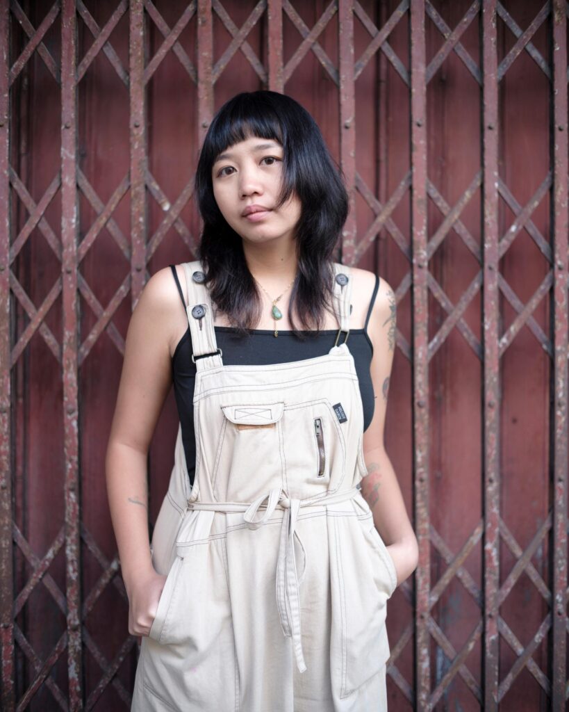 Member of Zua, Cinema and Chalan in beige overalls before a red folding gate in Bangkok.
