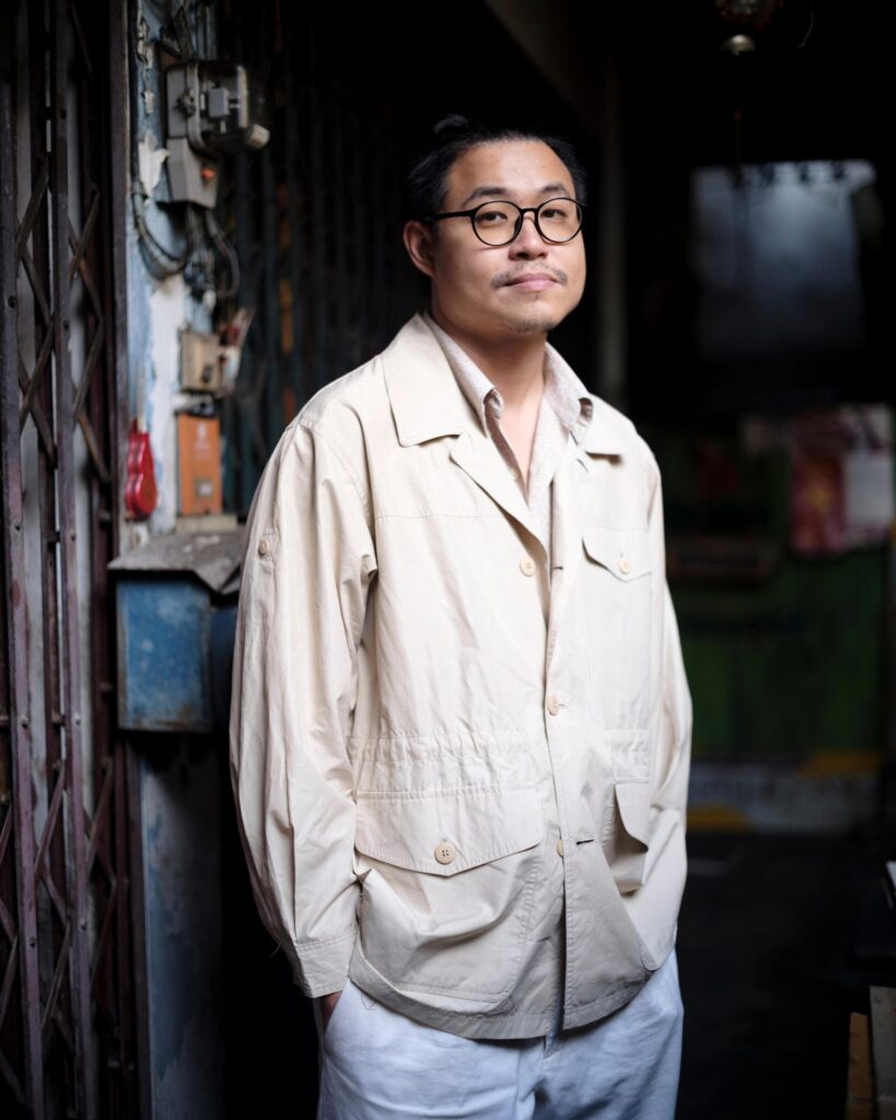 Member of Zua, Cinema and Chalan in glasses and a beige shirt in a Bangkok doorway.