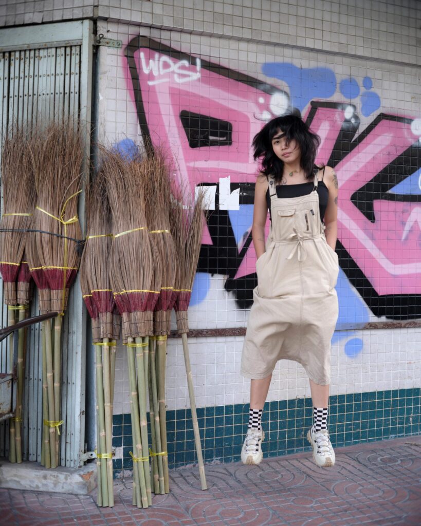Member of Zua, Cinema and Chalan jumping beside graffiti and bundled brooms in Bangkok.