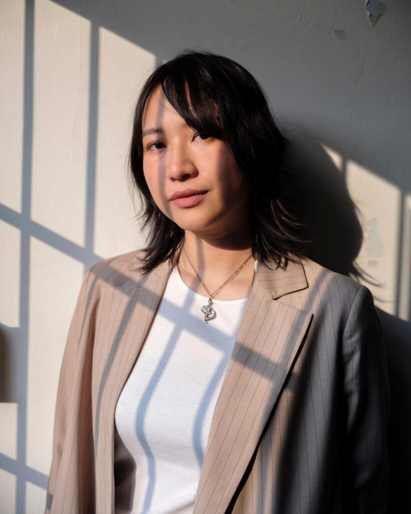 Member of Zua, Cinema and Chalan in a beige jacket, with window shadows across the wall in Bangkok.