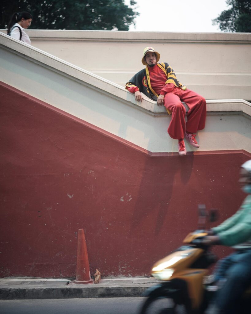 Banana Freeman sitting on a red-and-white stair wall as a motorcycle passes in Bangkok.