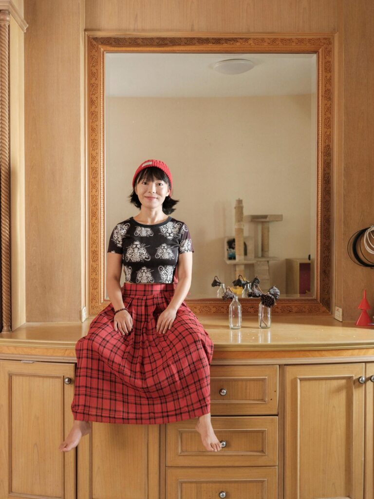 Chun Li in a red hat and plaid skirt, seated in front of a mirror at home in Bangkok.