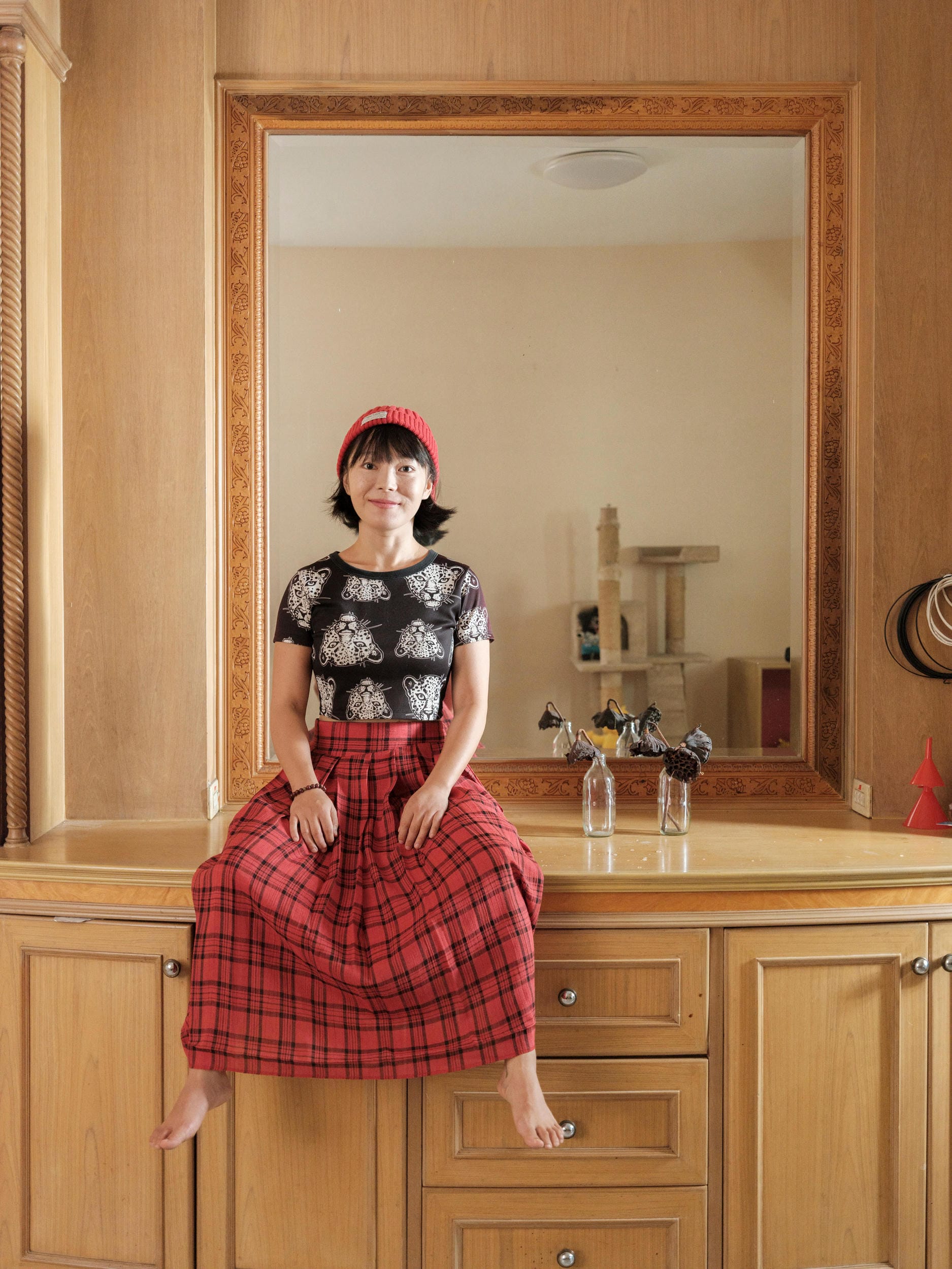Chun Li in a red hat and plaid skirt, seated in front of a mirror at home in Bangkok.