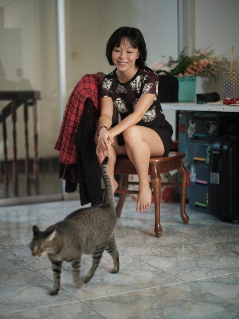 Chun Li seated on a wooden chair while a cat walks across the room in Bangkok.