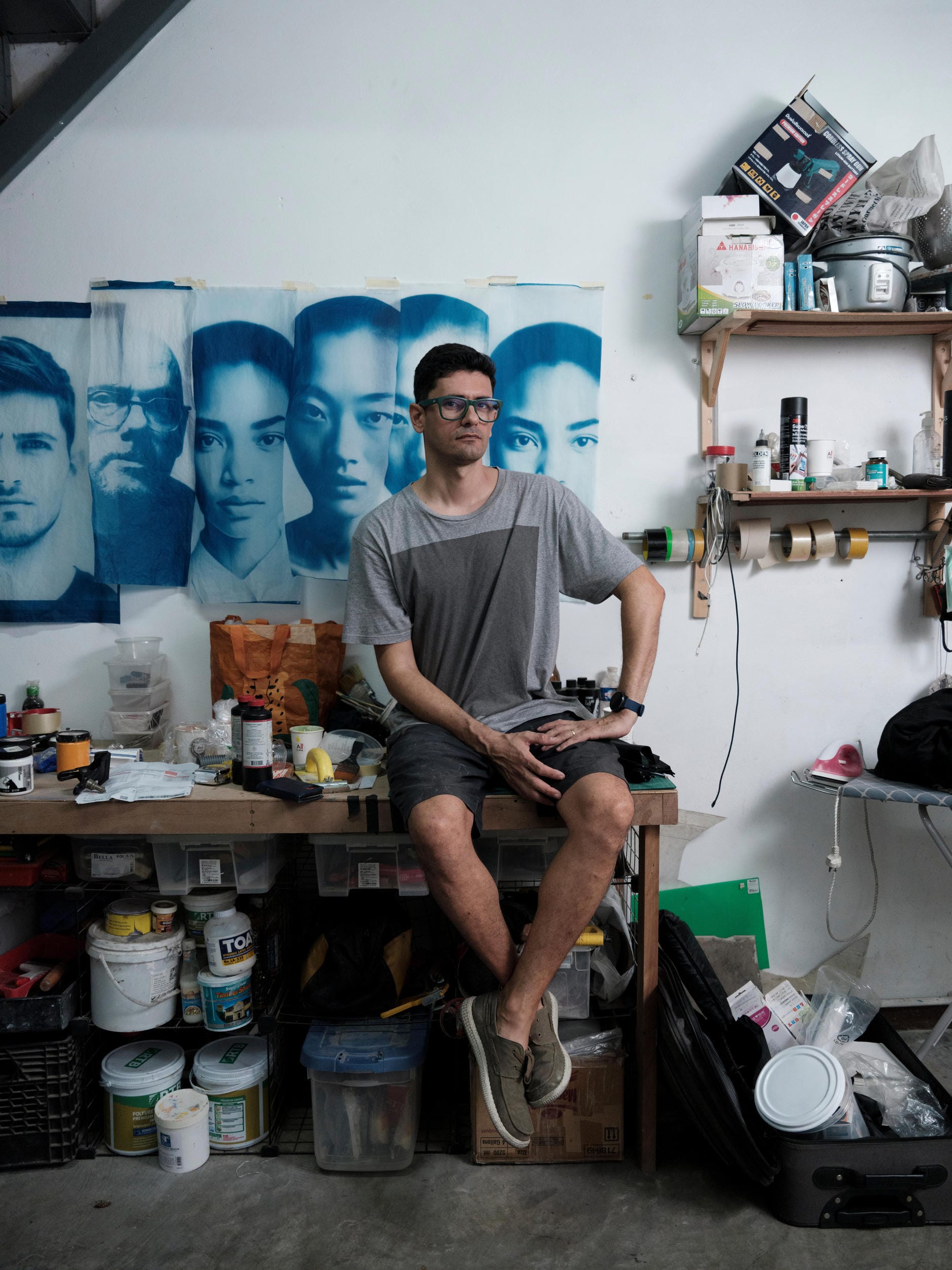 François Cambe seated at a studio workbench beneath blue portrait prints in Bangkok.