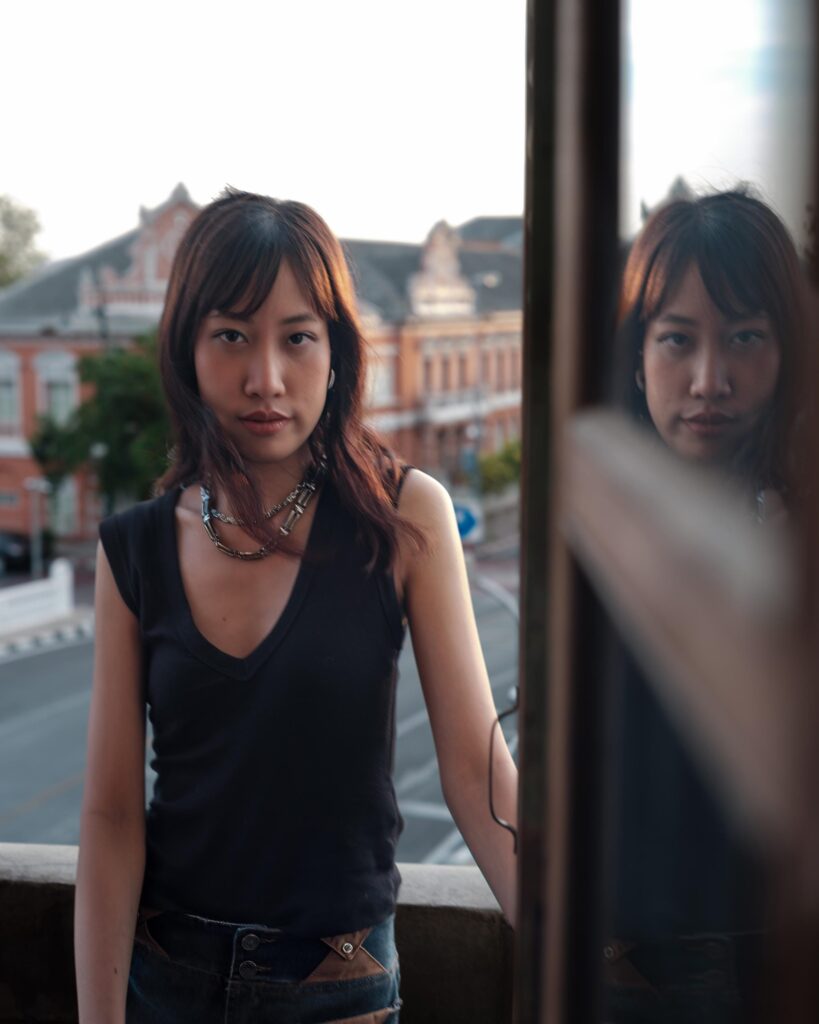 Issiree Shereen Sarntisart beside a window reflecting her face in Bangkok.