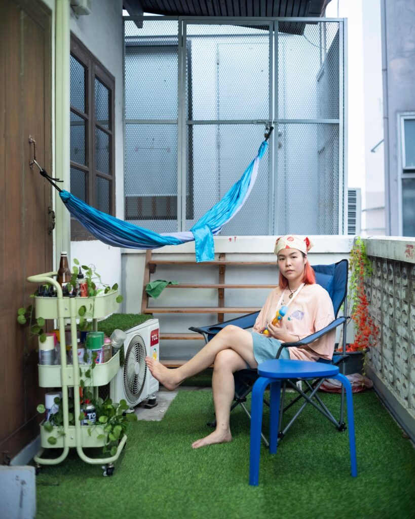 Jib Peerada (StupidShit) seated on a balcony with plants and a blue hammock in Bangkok.