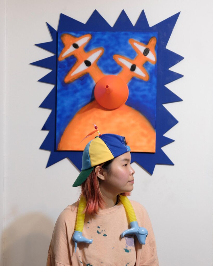 Jib Peerada (StupidShit) beneath a colourful artwork, wearing a toy-covered shirt in Bangkok.