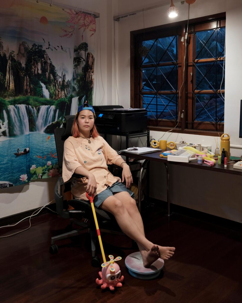 Jib Peerada (StupidShit) seated beside a desk and large artwork at home in Bangkok.
