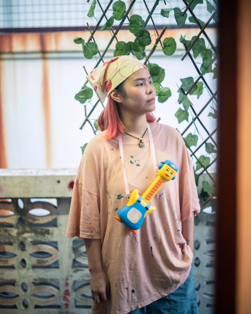 Jib Peerada (StupidShit) wearing a toy guitar necklace beside climbing plants in Bangkok.