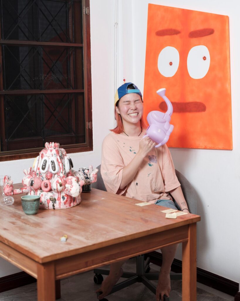 Jib Peerada (StupidShit) holding a pink sculpture at a table beside an orange artwork in Bangkok.
