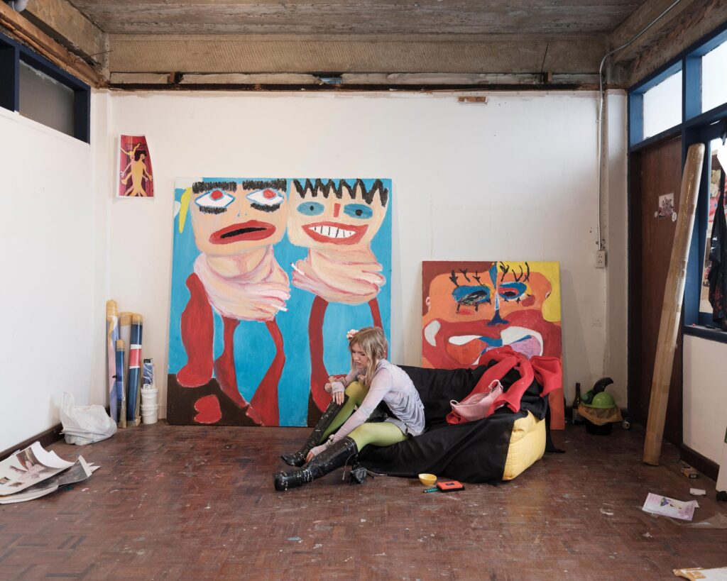 Mascha Serga seated on her studio floor beside large colourful paintings in Bangkok.