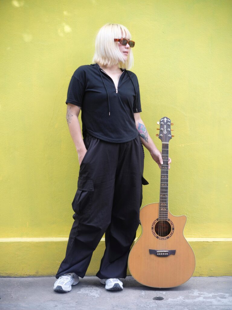 Nadia Mania standing with an acoustic guitar against a yellow wall in Chiang Mai.