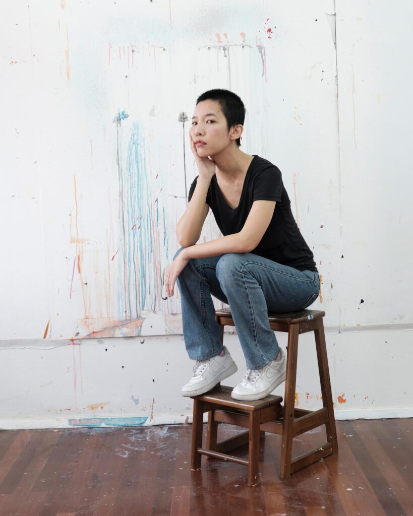 Pare Patcharapa Inchang seated on a wooden stool beside a painting in Chiang Mai.