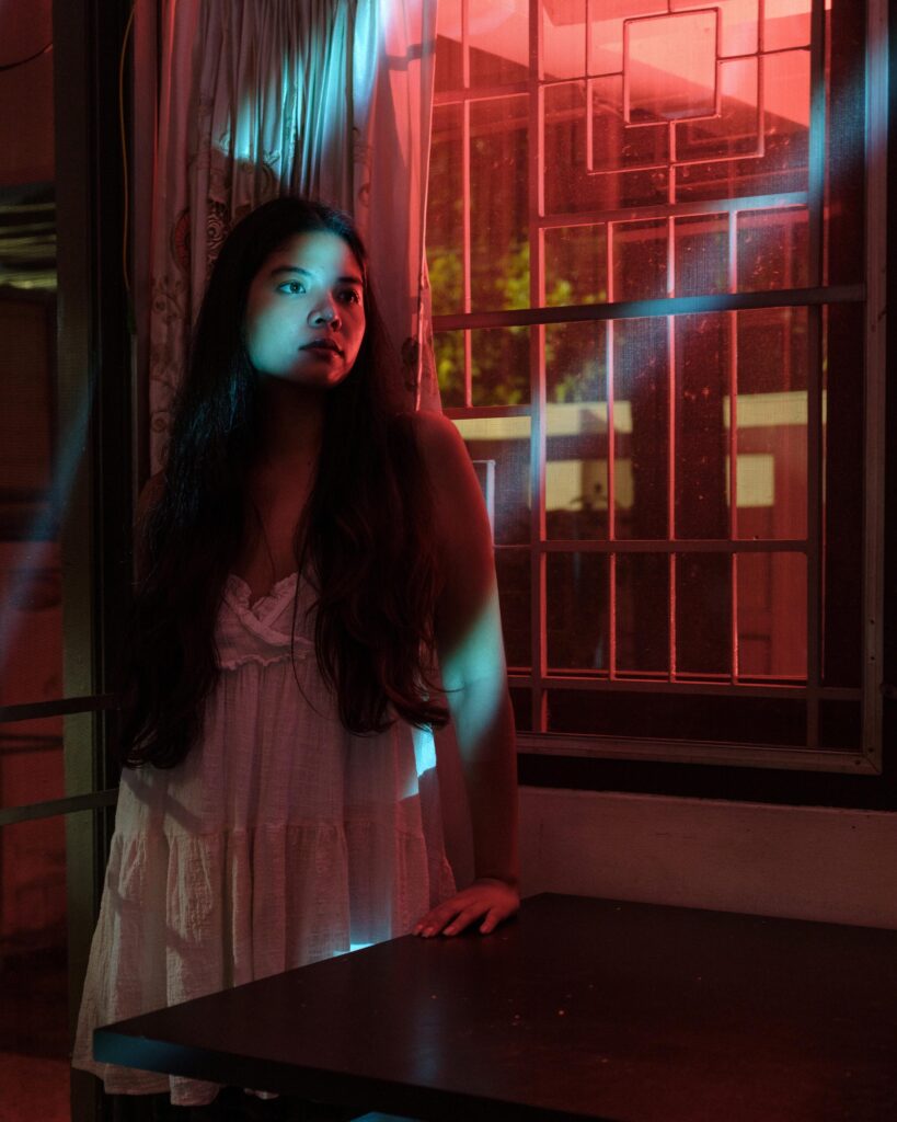 Pavee Vaidyanond beside a window illuminated by red and turquoise light at home in Bangkok.