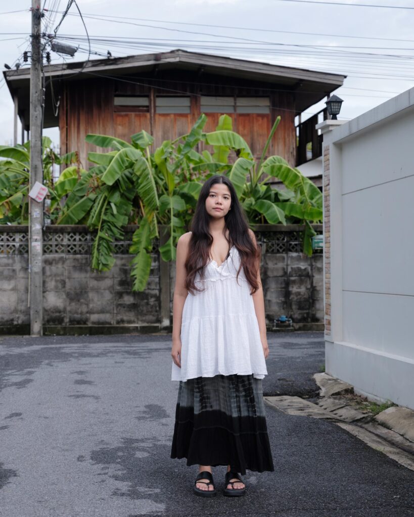 Pavee Vaidyanond standing in a Bangkok lane with plants and a wooden house behind her.