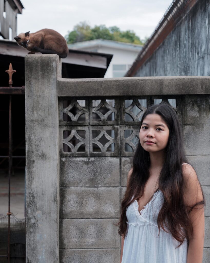 Pavee Vaidyanond beside a concrete wall with a cat resting above her in Bangkok.