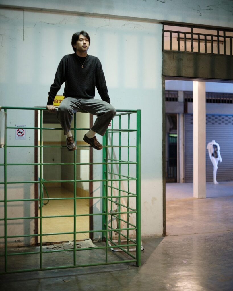 Peerapon Boonthep sitting on a green metal railing at Artcade Gallery in Phayao.