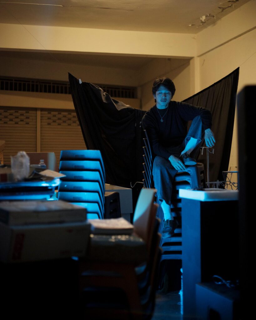 Peerapon Boonthep sitting above stacked chairs in blue light at Artcade Gallery in Phayao.