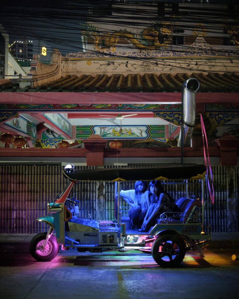 Phoenixe and Klein seated in a tuk-tuk under colourful light in Bangkok.
