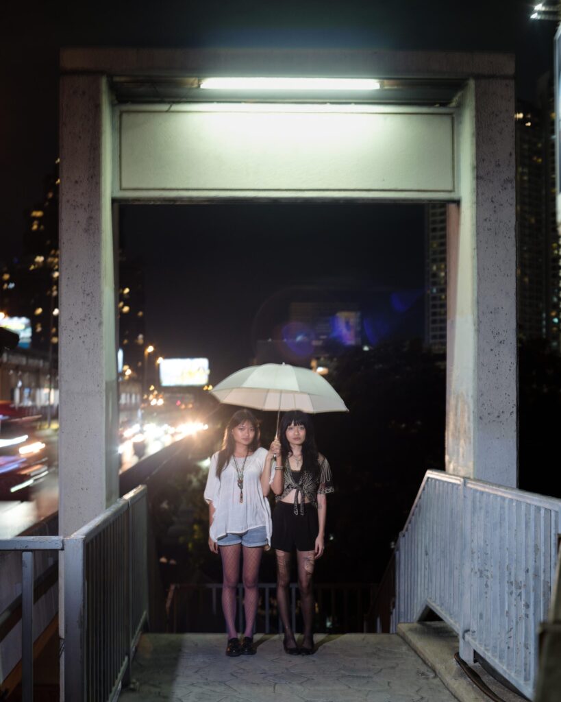 Phoenixe and Klein sharing an umbrella at the top of a Bangkok staircase at night.