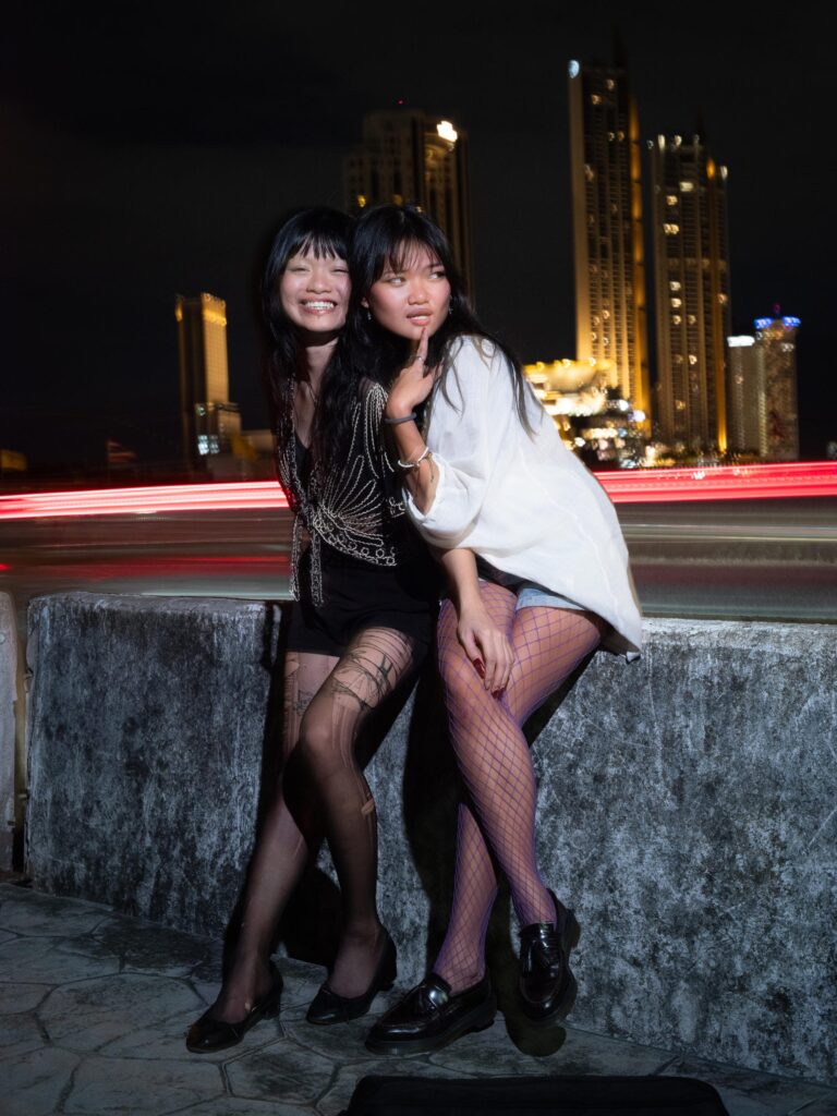 Phoenixe and Klein seated on a roadside wall with light trails behind them in Bangkok.