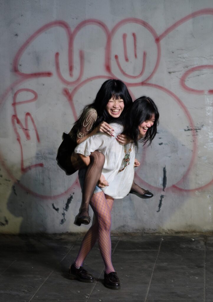 Phoenixe and Klein giving a piggyback ride in front of a graffiti-covered wall in Bangkok.
