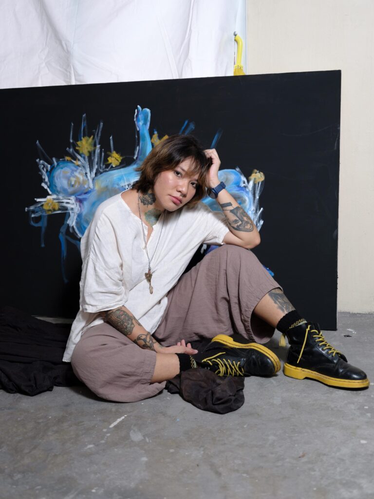 Ploypim sitting on the studio floor in front of a dark painting in Bangkok.