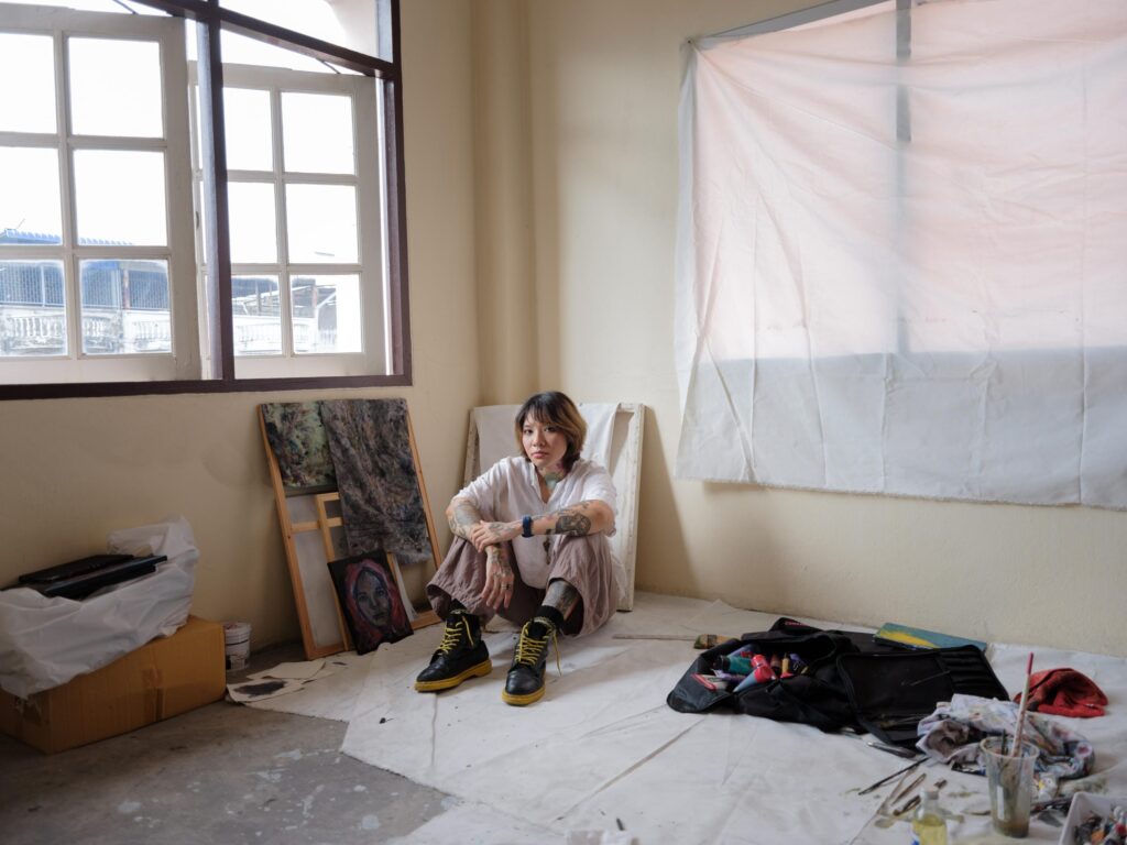 Ploypim seated in her Bangkok studio beside canvases and painting materials.