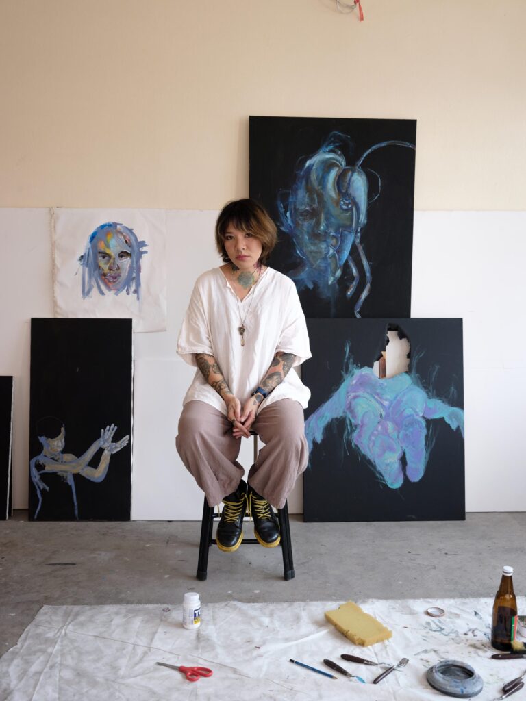 Ploypim seated on a stool in front of several paintings in her Bangkok studio.