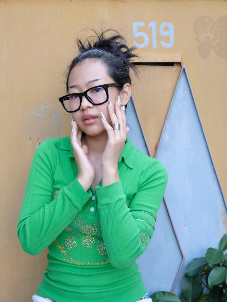Q3Q Community member wearing black glasses and a green top, with decorated nails against their cheeks in Bangkok.