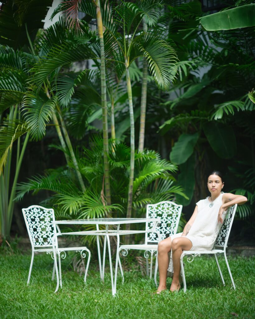 Rosalia Engchuan seated at a white garden table surrounded by tropical plants in Bangkok.