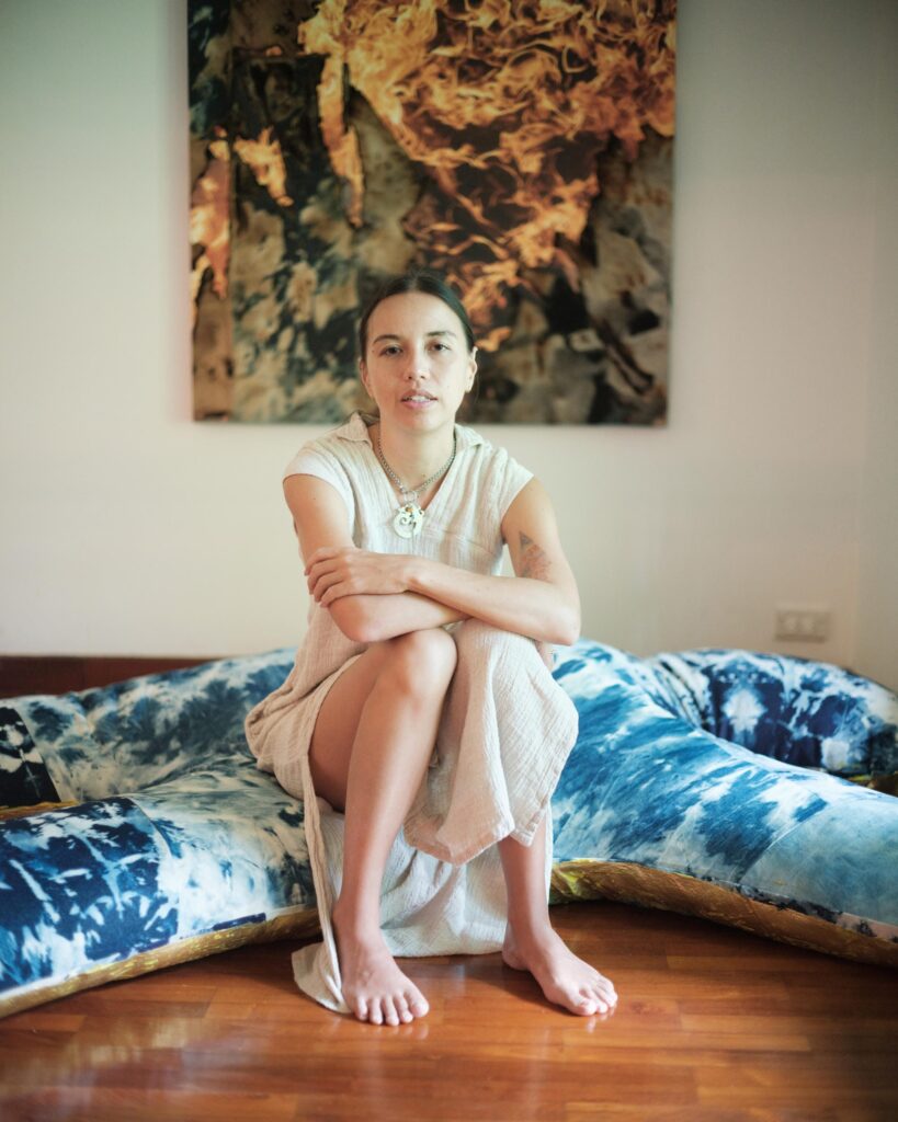 Rosalia Engchuan seated on a wooden floor beside blue cushions and an artwork in Bangkok.