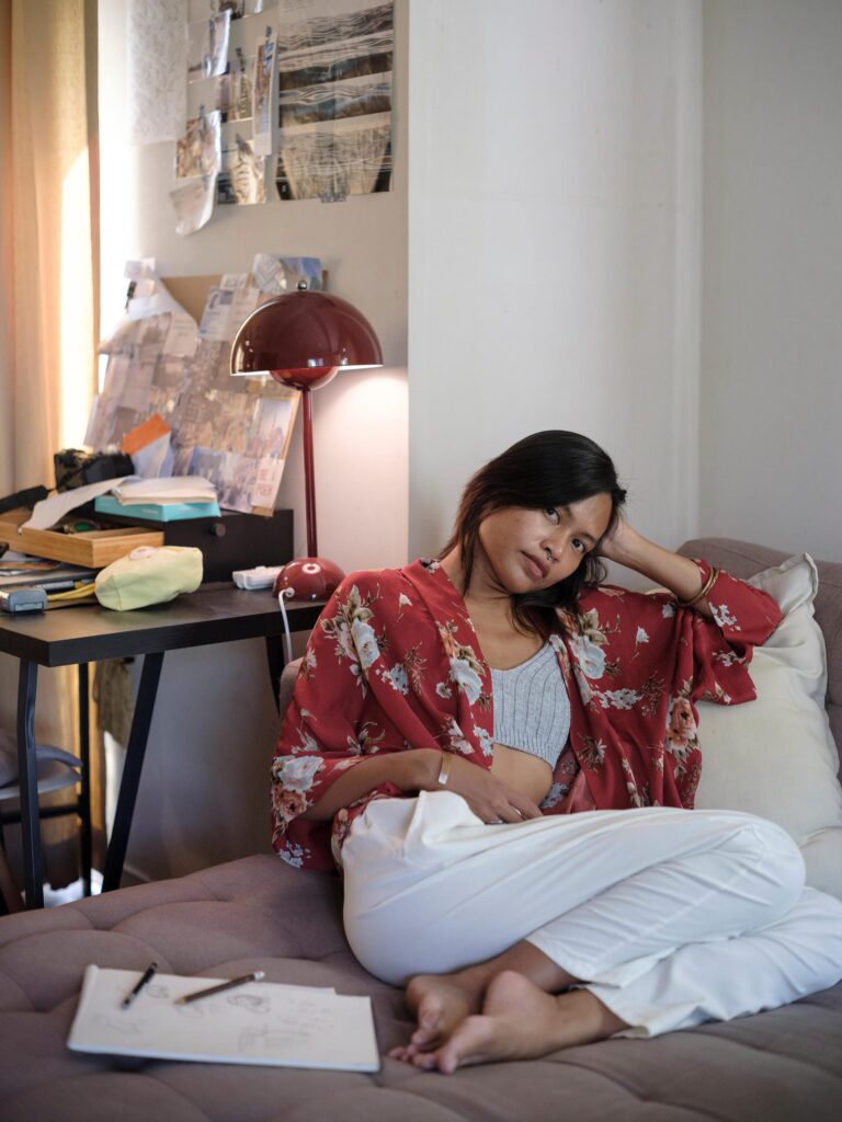 Sacha Piisasha reclining on a sofa beside books and a red lamp in Nonthaburi.