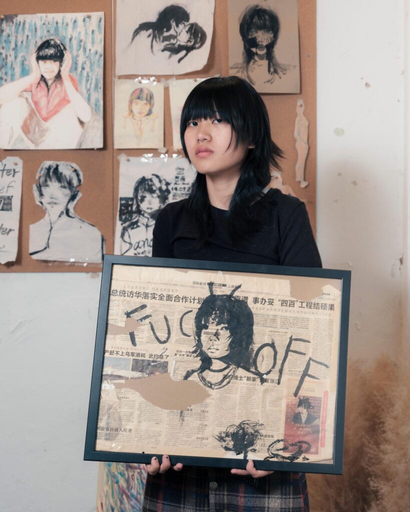 Sandee holding a framed artwork in front of a wall of portraits in Bangkok.