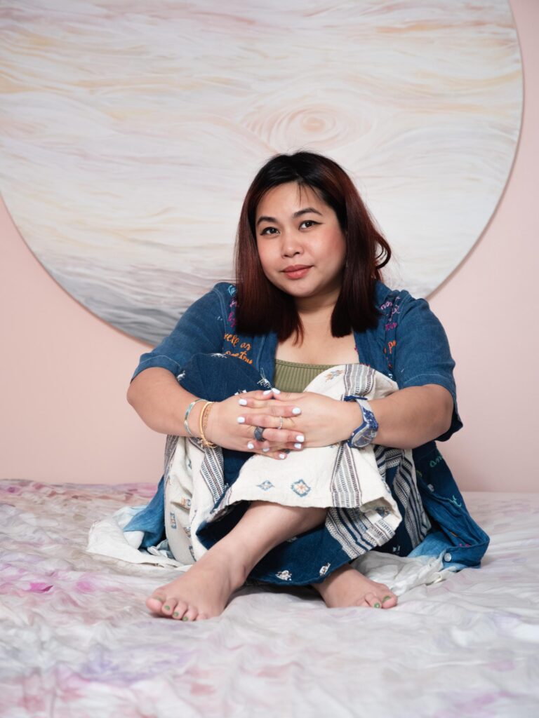 Tamruja Dharmasaroja seated on a bed beneath a round painting at home in Bangkok.