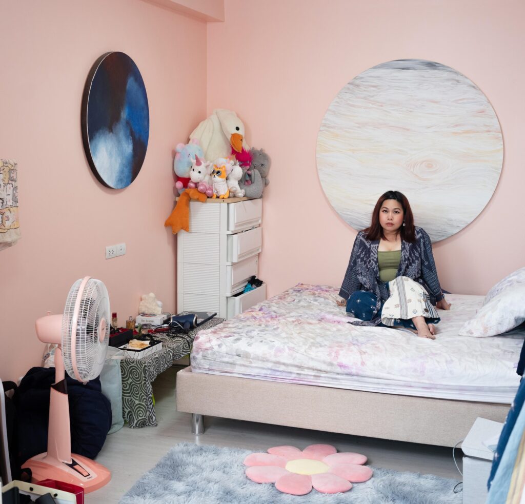 Tamruja Dharmasaroja seated in a pink bedroom with artworks and toys in Bangkok.