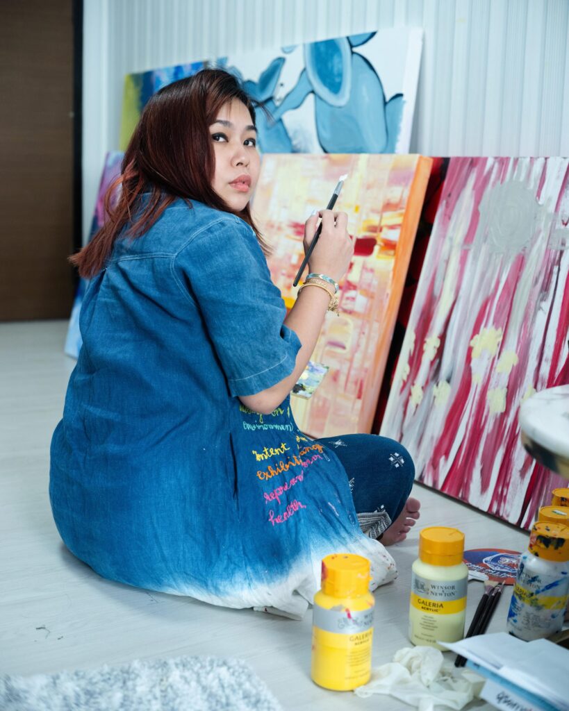 Tamruja Dharmasaroja seated on the floor painting a colourful canvas in Bangkok.