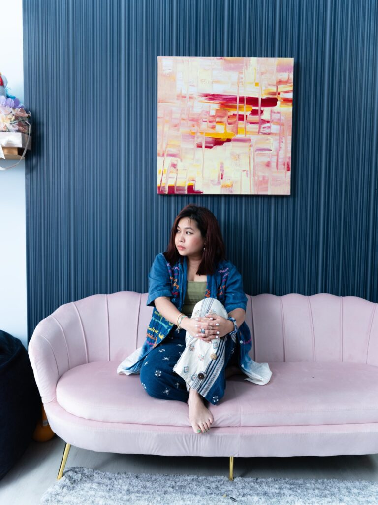 Tamruja Dharmasaroja seated on a pink sofa beneath an abstract painting in Bangkok.